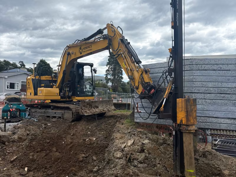 Excavator attachment working on a vertical retaining post beside installed retaining panels