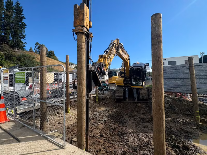 Excavator-mounted piling attachment driving a large timber pile on a construction site