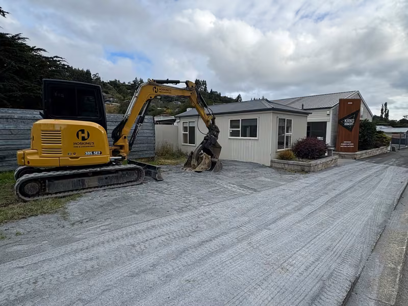 Excavator carrying out earthworks on a prepared gravel area beside an existing building